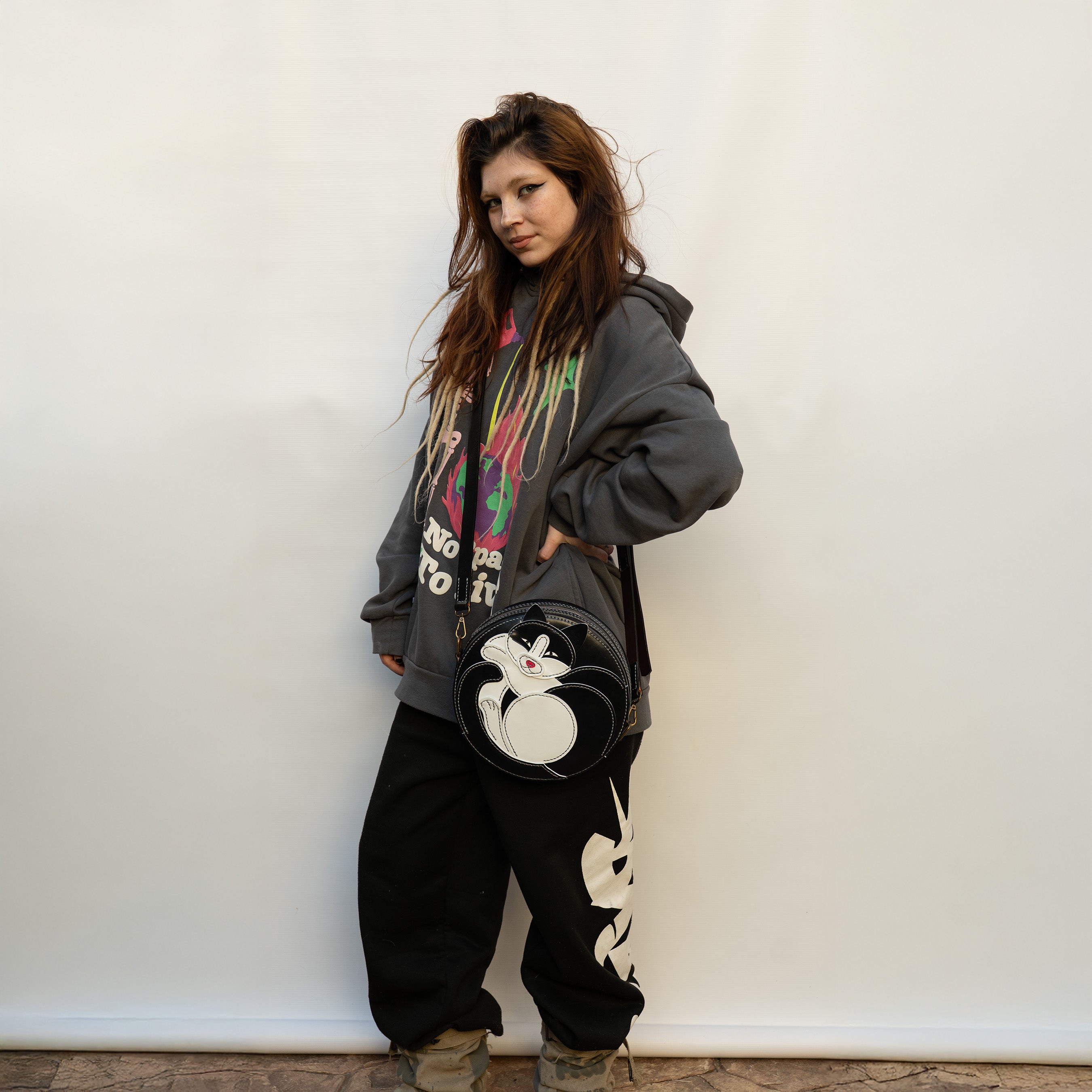 Person wearing a gray hoodie with colorful graphics and black pants, holding a black bag with a white cat  design against a white wall.