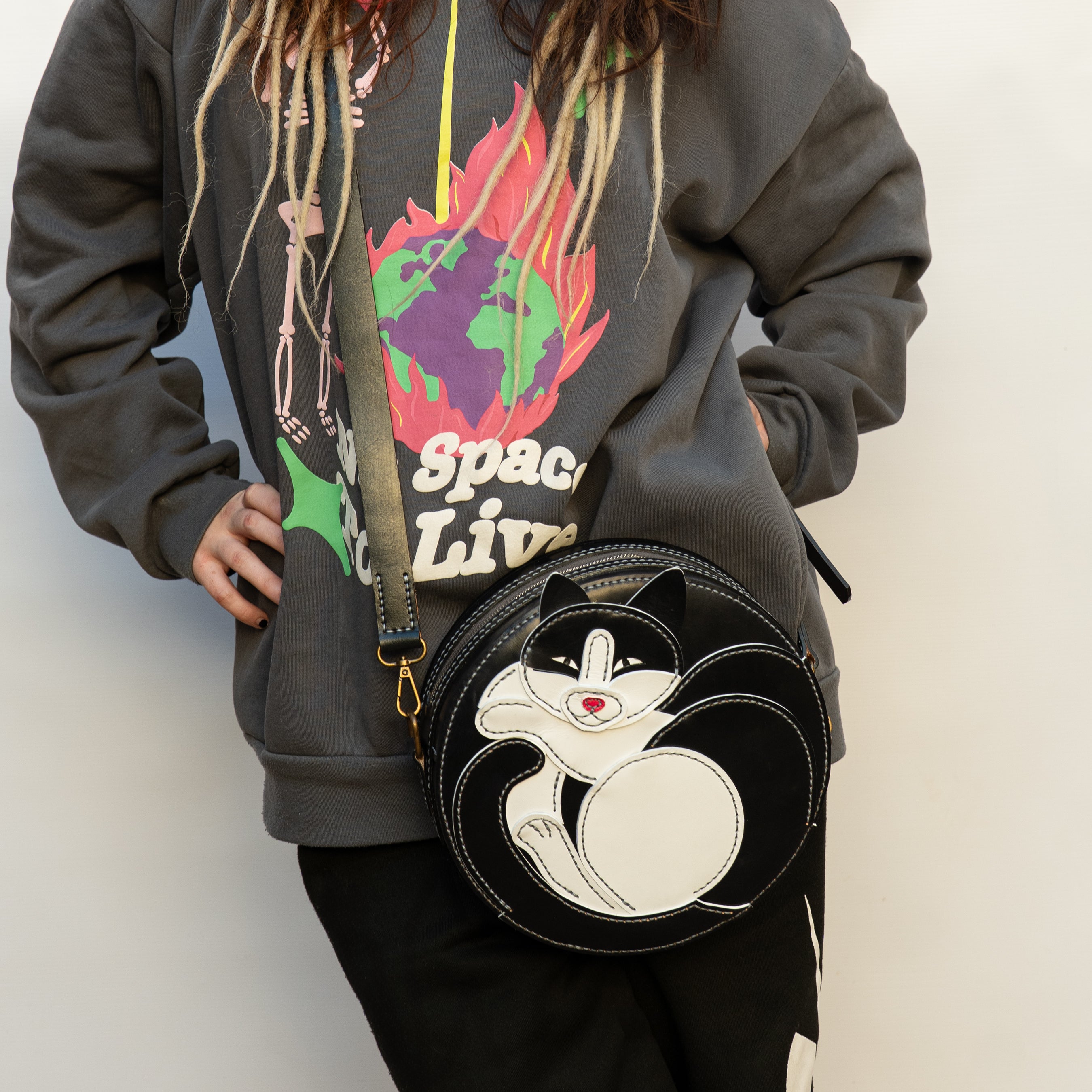 Person wearing a hoodie with a colorful design, holding a black round bag with a cat aplique.