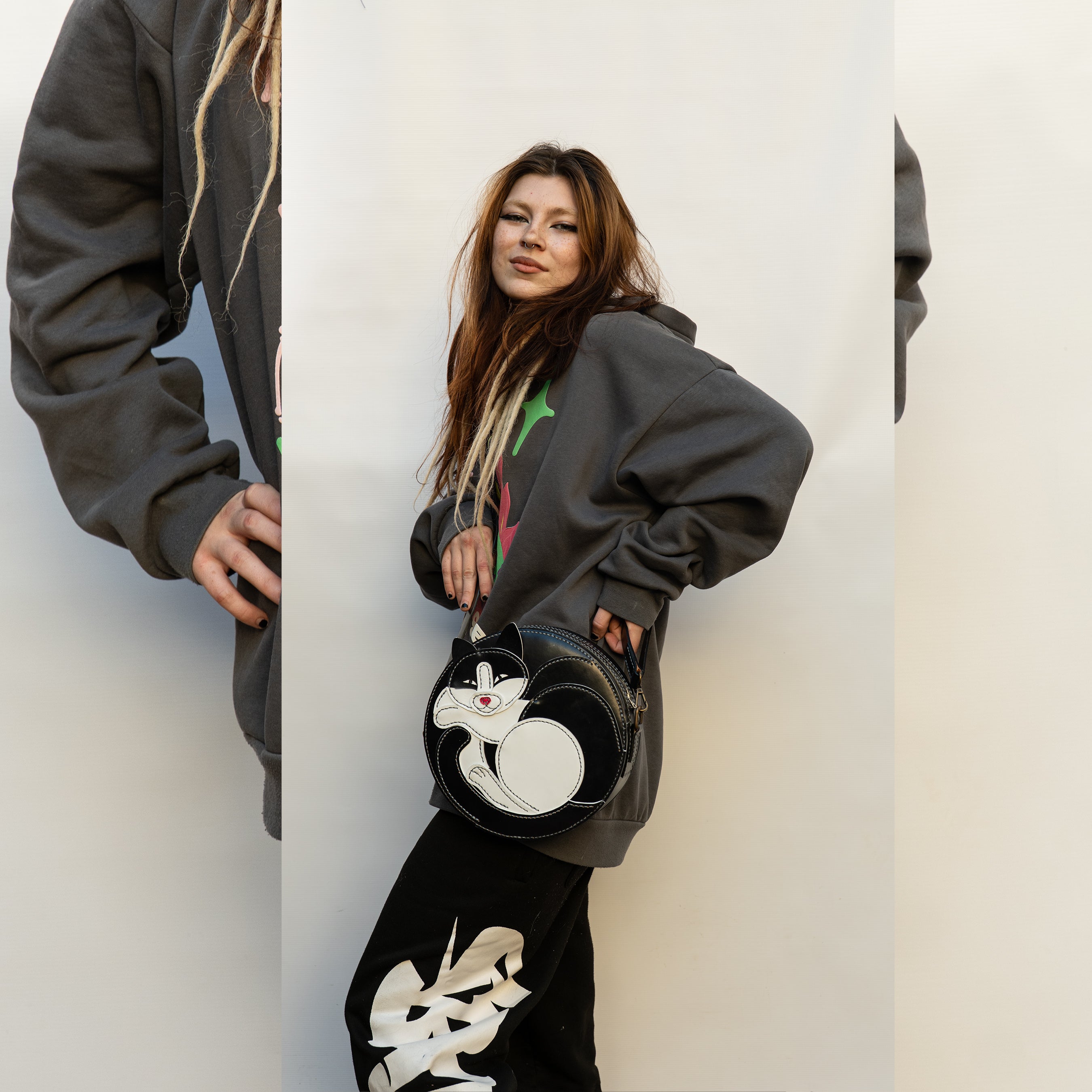 Person wearing a dark hoodie and black pants with white designs, holding a black bag with a cat design.