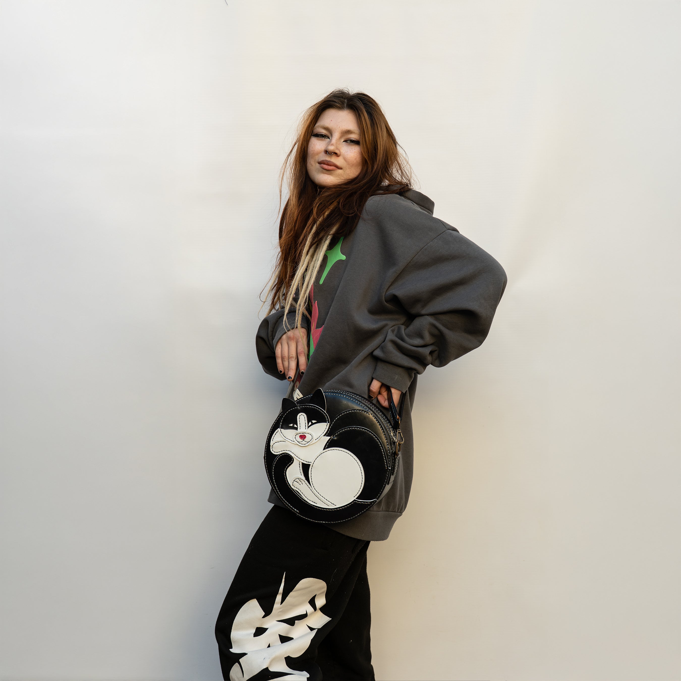 Person wearing a gray hoodie and black pants with white designs, holding a black bag with a cat design.